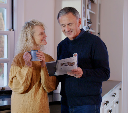 A married couple browsing a magazine in their kitchen. The woman is holding a mug and gazing at her husband.